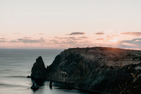 A red burning sunset over the sea with rocky volcanic cliff. Abstract nature summer ocean sea background. Small waves on golden warm water surface with bokeh lights from sun.の写真素材