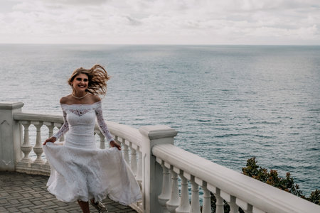 Romantic bride, blond girl in white wedding dress with open shoulders posing on open terrace with backdrop of the sea and rocks. Stylish young woman standing on terrace and looking on ocean at sunsetの写真素材