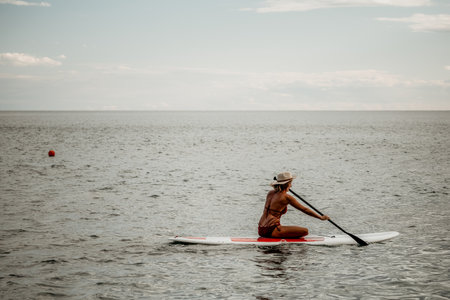 Woman in red bikini on sup board. Happy lady with blond hair in red bathing suit chilling and sunbathing by turquoise sea ocean on sunset. Holiday, vacation and recreational concept.の写真素材