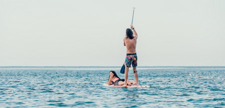 Sea woman and man on sup. Silhouette of happy young woman and man, surfing on SUP board, confident paddling through water surface. Idyllic sunset. Active lifestyle at sea or river.の写真素材