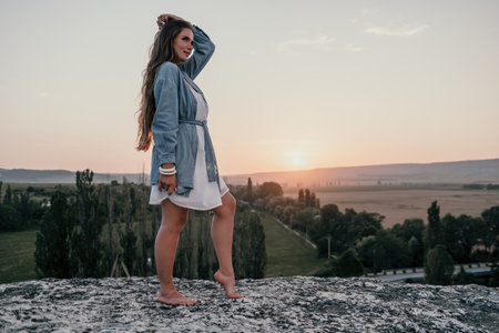 Happy woman standing with her back on the sunset in nature in summer with open hands. Romantic beautiful bride in white boho dress posing with mountains on sunsetの写真素材