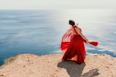 Woman in red dress on sea. Side view a Young beautiful sensual woman in a red long dress posing on a rock high above the sea on sunset. Girl on the nature on blue sky background. Fashion photo.の写真素材