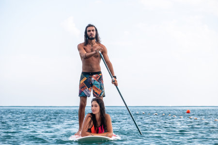 Sea woman and man on sup. Silhouette of happy young woman and man, surfing on SUP board, confident paddling through water surface. Idyllic sunset. Active lifestyle at sea or river.の写真素材