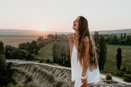 Happy woman standing with her back on the sunset in nature in summer with open hands. Romantic beautiful bride in white boho dress posing with mountains on sunsetの写真素材