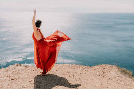 Woman in red dress on sea. Side view a Young beautiful sensual woman in a red long dress posing on a rock high above the sea on sunset. Girl on the nature on blue sky background. Fashion photo.の写真素材