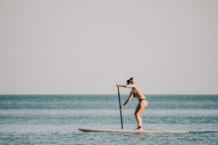 Sea woman sup. Silhouette of happy middle aged tanned woman in rainbow bikini, surfing on SUP board, confident paddling through water surface. Idyllic sunset. Active lifestyle at sea or river.の写真素材