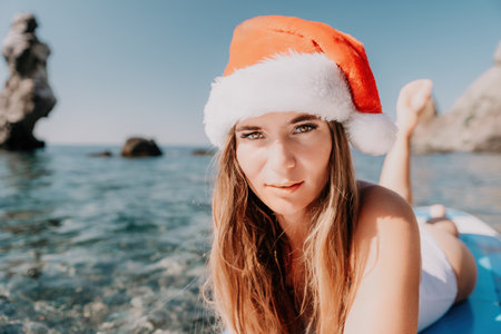 Woman sea sup. Close up portrait of happy young caucasian woman with long hair in Santa hat looking at camera and smiling. Cute woman portrait in a white bikini posing on sup board in the seaの写真素材