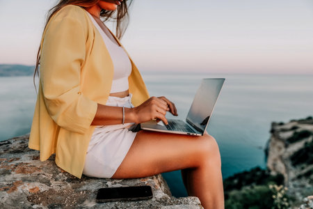 Digital nomad, Business woman working on laptop by the sea. Pretty lady typing on computer by the sea at sunset, makes a business transaction online from a distance. Freelance, remote work on vacationの写真素材