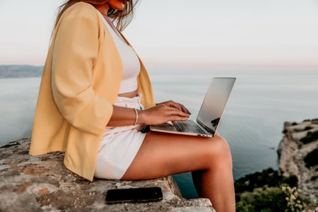 Digital nomad, Business woman working on laptop by the sea. Pretty lady typing on computer by the sea at sunset, makes a business transaction online from a distance. Freelance, remote work on vacationの写真素材