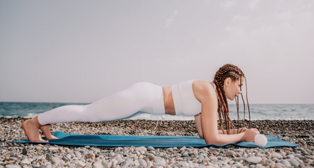 Woman sea yoga. Well looking middle aged woman with braids dreadlocks in white leggings and tops doing stretching pilates on yoga mat near sea. Female fitness yoga routine concept. Healthy lifestyle.の写真素材