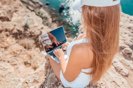 Digital nomad, woman in the hat, a business woman with a laptop sits on the rocks by the sea during sunset, makes a business transaction online from a distance. Freelance, remote work on vacation.の写真素材