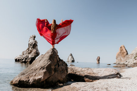 Woman travel sea. Young Happy woman in a long red dress posing on a beach near the sea on background of volcanic rocks, like in Iceland, sharing travel adventure journeyの写真素材