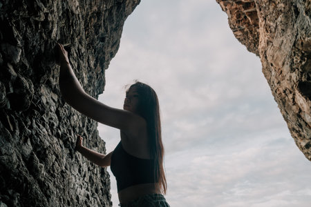 Sports Woman Climbing The Rock. Young woman With slim fit body climbing in volcanic basalt cave with beautiful sea view. The athlete girl trains in nature. Woman overcomes difficult climbing route.の写真素材