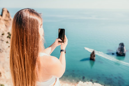 Digital nomad, woman in the hat, a business woman with a laptop sits on the rocks by the sea during sunset, makes a business transaction online from a distance. Freelance, remote work on vacation.の写真素材