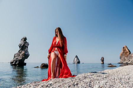 Woman travel sea. Happy tourist in red dress enjoy taking picture outdoors for memories. Woman traveler posing on the rock at sea bay surrounded by volcanic mountains, sharing travel adventure journeyの写真素材