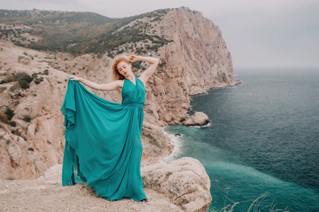 Redhead woman portrait. Curly redhead young caucasian woman with freckles looking at camera and smiling. Close up portrait cute woman in a mint long dress posing on a volcanic rock high above the seaの写真素材