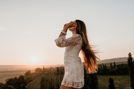 Happy woman in white boho dress on sunset in mountains. Romantic woman with long hair standing with her back on the sunset in nature in summer with open hands. Silhouette. Nature. Sunset.の写真素材