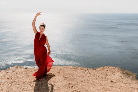 Woman in red dress on sea. Side view a Young beautiful sensual woman in a red long dress posing on a rock high above the sea on sunset. Girl on the nature on blue sky background. Fashion photo.の写真素材