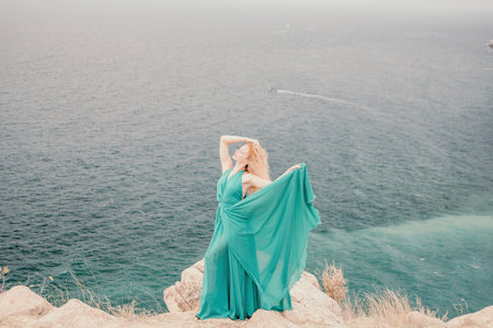 Redhead woman portrait. Curly redhead young caucasian woman with freckles looking at camera and smiling. Close up portrait cute woman in a mint long dress posing on a volcanic rock high above the seaの写真素材