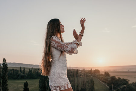 Happy woman in white boho dress on sunset in mountains. Romantic woman with long hair standing with her back on the sunset in nature in summer with open hands. Silhouette. Nature. Sunset.の写真素材
