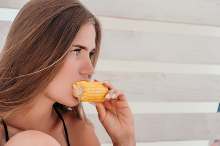 Woman eats corn on beach. Vegetarian hipster woman eat fresh organic grilled corn. Happy lady on sea beach sunset or ocean sunrise. Travel, explore, active yoga and meditation lifestyle concept.の写真素材