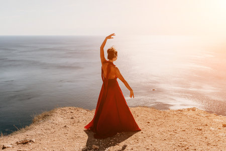 Woman in red dress on sea. Side view a Young beautiful sensual woman in a red long dress posing on a rock high above the sea on sunset. Girl on the nature on blue sky background. Fashion photo.の写真素材