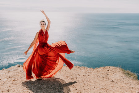 Woman in red dress on sea. Side view a Young beautiful sensual woman in a red long dress posing on a rock high above the sea on sunset. Girl on the nature on blue sky background. Fashion photo.の写真素材
