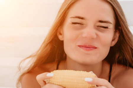 Woman eats corn on beach. Vegetarian hipster woman eat fresh organic grilled corn. Happy lady on sea beach sunset or ocean sunrise. Travel, explore, active yoga and meditation lifestyle concept.の写真素材