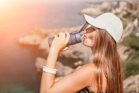 Woman travel sea. Happy tourist drink water on hot summer day. Woman traveler looks at the edge of the cliff on the sea bay of mountains, sharing travel adventure journeyの写真素材