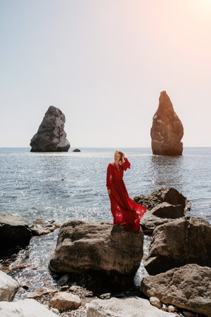 Woman summer travel sea. Happy tourist in long red dress enjoy taking picture outdoors for memories. Woman traveler posing on beach at sea surrounded by volcanic mountains, sharing travel adventureの写真素材