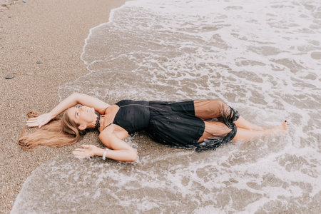 Woman summer travel sea. Happy tourist in black dress enjoy taking picture outdoors for memories. Woman traveler posing on sea beach surrounded by volcanic mountains, sharing travel adventure journeyの写真素材