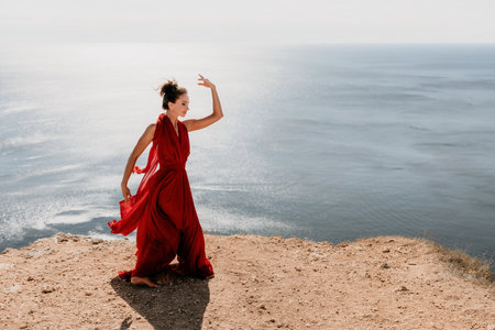 Woman in red dress on sea. Side view a Young beautiful sensual woman in a red long dress posing on a rock high above the sea on sunset. Girl on the nature on blue sky background. Fashion photo.の写真素材