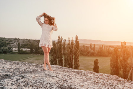 Happy woman in white boho dress on sunset in mountains. Romantic woman with long hair standing with her back on the sunset in nature in summer with open hands. Silhouette. Nature. Sunset.の写真素材