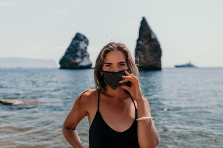 Woman summer travel sea. Happy tourist in black mask enjoy taking picture outdoors for memories. Woman traveler posing on the beach at sea surrounded by volcanic mountains, sharing travel adventureの写真素材