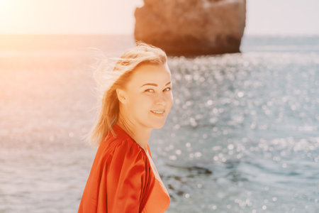 Woman summer travel sea. Happy tourist in long red dress enjoy taking picture outdoors for memories. Woman traveler posing on beach at sea surrounded by volcanic mountains, sharing travel adventureの写真素材