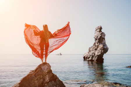 Woman travel sea. Young Happy woman in a long red dress posing on a beach near the sea on background of volcanic rocks, like in Iceland, sharing travel adventure journeyの写真素材