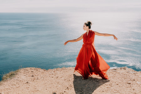 Woman in red dress on sea. Side view a Young beautiful sensual woman in a red long dress posing on a rock high above the sea on sunset. Girl on the nature on blue sky background. Fashion photo.の写真素材