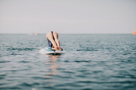 Woman sup yoga. Middle age sporty woman practising yoga pilates on paddle sup surfboard. Female stretching doing workout on sea water. Modern individual female hipster outdoor summer sport activity.の写真素材