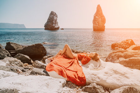 Woman summer travel sea. Happy tourist in long red dress enjoy taking picture outdoors for memories. Woman traveler posing on beach at sea surrounded by volcanic mountains, sharing travel adventureの写真素材