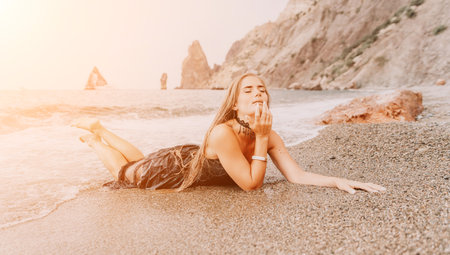 Woman summer travel sea. Happy tourist in black dress enjoy taking picture outdoors for memories. Woman traveler posing on sea beach surrounded by volcanic mountains, sharing travel adventure journeyの写真素材