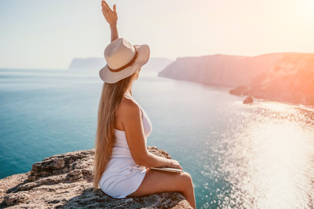 Digital nomad, woman in the hat, a business woman with a laptop sits on the rocks by the sea during sunset, makes a business transaction online from a distance. Freelance, remote work on vacation.の写真素材