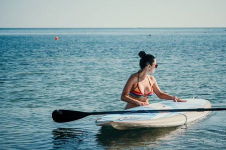 Sea woman sup. Silhouette of happy middle aged tanned woman in rainbow bikini, surfing on SUP board, confident paddling through water surface. Idyllic sunset. Active lifestyle at sea or river.の写真素材