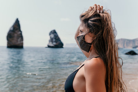Woman summer travel sea. Happy tourist in black mask enjoy taking picture outdoors for memories. Woman traveler posing on the beach at sea surrounded by volcanic mountains, sharing travel adventureの写真素材
