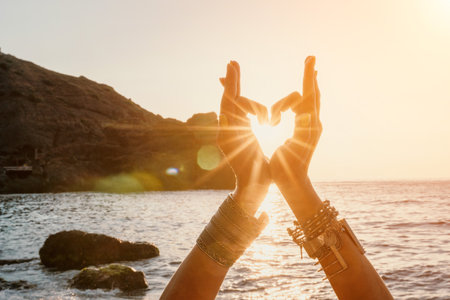Woman sea yoga. Happy woman in a white swimsuit and boho-style bracelets making a heart shape with her hands while practicing yoga on a mat by sea at sunset. Healthy lifestyle, harmony and meditationの写真素材