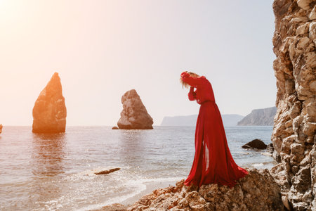 Woman summer travel sea. Happy tourist in long red dress enjoy taking picture outdoors for memories. Woman traveler posing on beach at sea surrounded by volcanic mountains, sharing travel adventureの写真素材