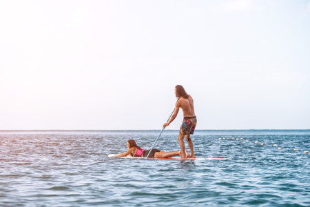Sea woman and man on sup. Silhouette of happy young woman and man, surfing on SUP board, confident paddling through water surface. Idyllic sunset. Active lifestyle at sea or river.の写真素材