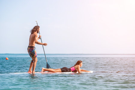 Sea woman and man on sup. Silhouette of happy young woman and man, surfing on SUP board, confident paddling through water surface. Idyllic sunset. Active lifestyle at sea or river. Slow motionの写真素材