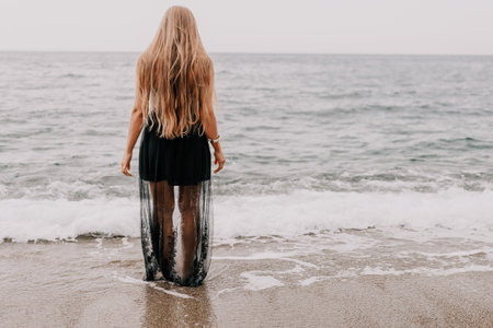 Woman summer travel sea. Happy tourist in black dress enjoy taking picture outdoors for memories. Woman traveler posing on sea beach surrounded by volcanic mountains, sharing travel adventure journeyの写真素材