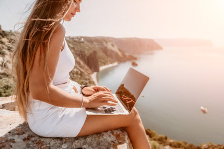Digital nomad, Business woman working on laptop by the sea. Pretty lady typing on computer by the sea at sunset, makes a business transaction online from a distance. Freelance, remote work on vacationの写真素材