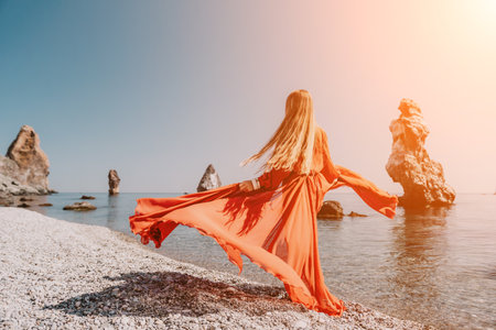 Woman travel sea. Happy tourist in red dress enjoy taking picture outdoors for memories. Woman traveler posing on the rock at sea bay surrounded by volcanic mountains, sharing travel adventure journeyの写真素材